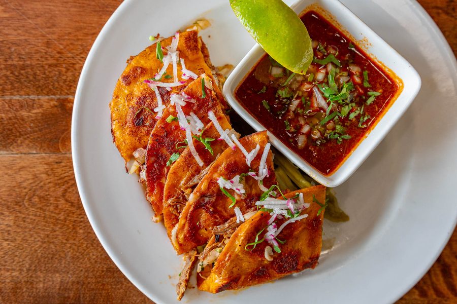Crisp red birria tacos topped with white onion and cilantro, with a cup of consommé and a lime wedge, at 5 Rabanitos