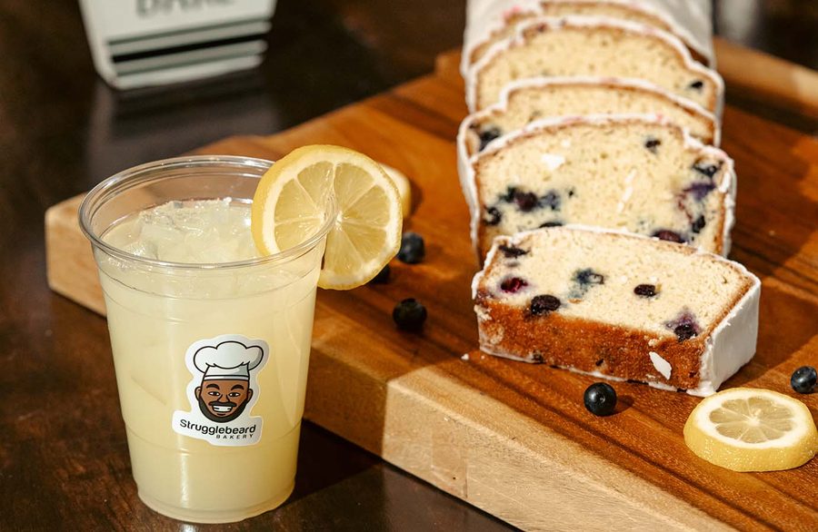 An iced lemonade in a StruggleBeard cup beside a sliced lemon blueberry loaf on a wooden board